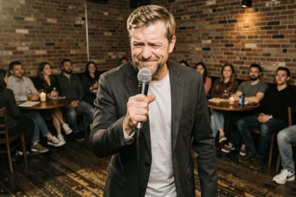 Humoriste sur scène dans un club de stand-up tenant un micro, expression ambiguë face au public, ambiance de soirée privée banalisée